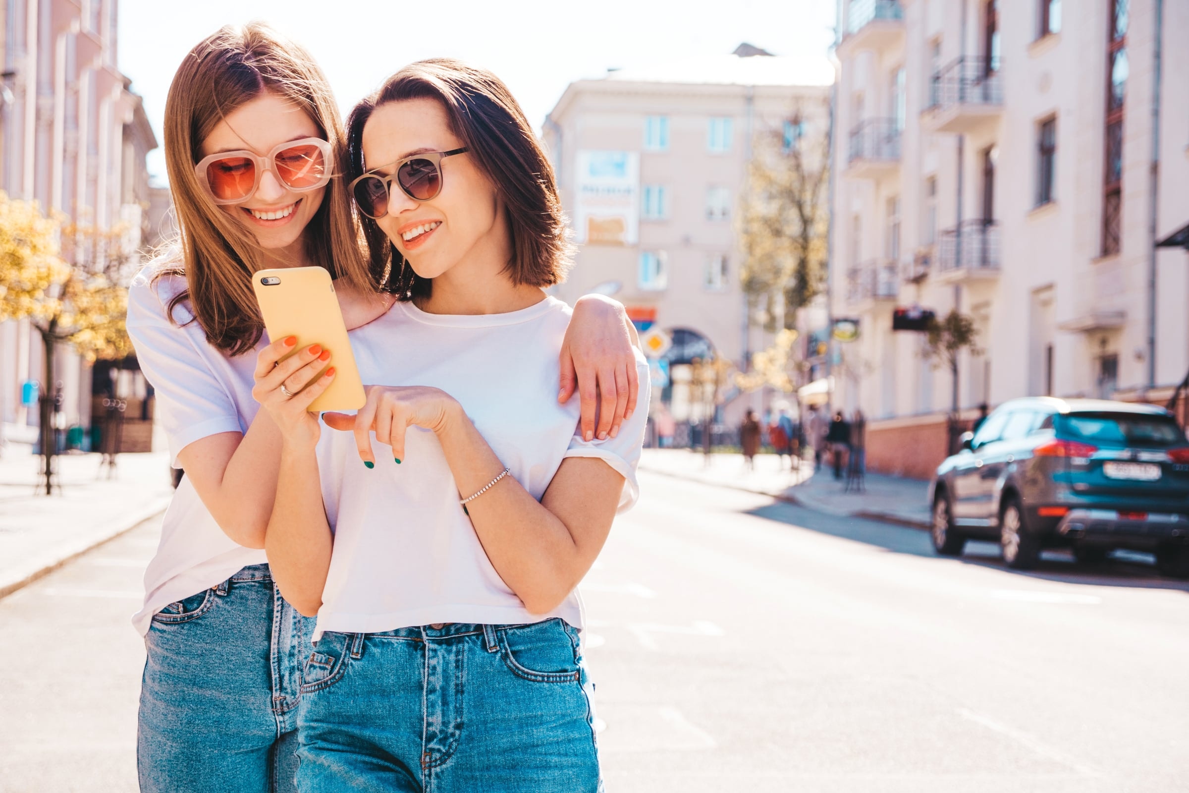 Two young friends in sunglasses smiling at a phone on a sunny street